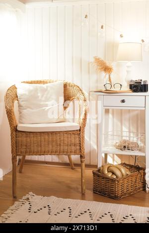 Wicker peacock chair with pillow and bedside table with book, retro camera, glasses and lamp in beige light wooden interior. Stock Photo