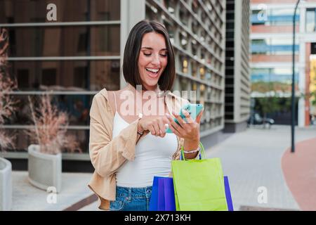 Smiling female carrying shopping bags and having fun watching a sale on a mobile phone. Happy caucasian lady browsing. Young pretty woman using a cellphone to purchase online on a market place app. High quality photo Stock Photo