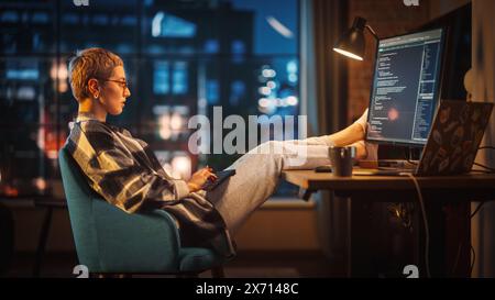 Young Woman Writing Code on Computer in Stylish Loft Apartment in the Evening. Creative Female Wearing Cozy Clothes, Resting Feet on Table and Using Wireless Keyboard. Urban City View from Big Window. Stock Photo