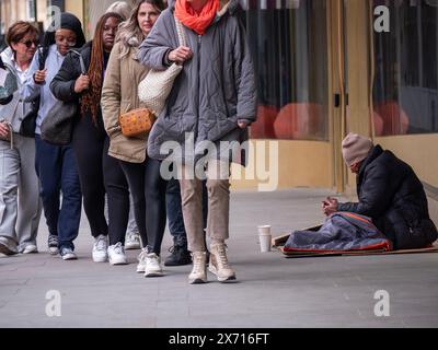 Homeless man in sleeping bag using mobile phone amongst crowds of shoppers,  Charing Cross Road London Stock Photo