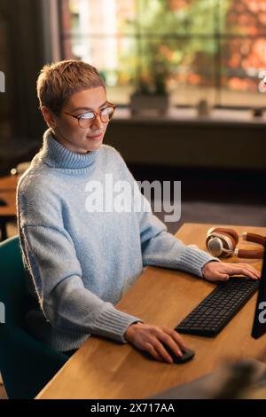 Vertical Screen: Young Attractive Woman Using Desktop Computer in Stylish Loft Apartment During the Day. Creative Female Checking Social Media, Browsing Internet, Wearing Cozy Clothes. Stock Photo
