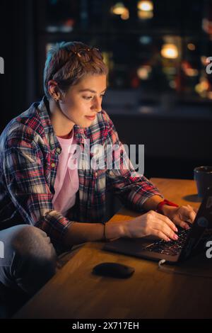 Vertical Screen: Young Attractive Woman Using Laptop Computer in Stylish Loft Apartment in the Evening. Creative Female Checking Social Media, Browsing Internet, Wearing Cozy Clothes. Stock Photo