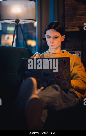 Vertical Screen: Young Handsome Man Working from Home on Laptop Computer in Stylish Loft Apartment in the Evening. Creative Person Checking Social Media, Browsing Internet. Stock Photo
