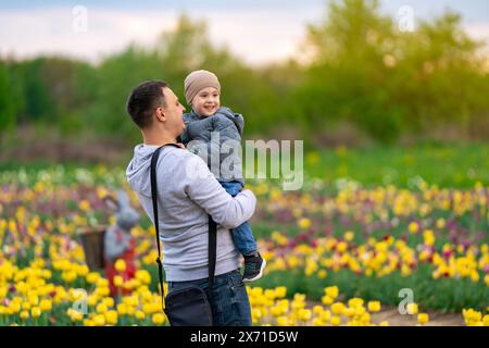 A happy father holds his smiling three-year-old son in hands in a tulip field on a spring day. Family time and leisure Stock Photo