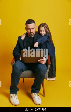 Father working online and his little daughter dressed formal attire pointing to screen against sunny-yellow background. Stock Photo