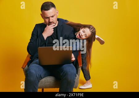Thoughtful father using laptop and his beloved, little daughter have fun while he works against sunny-yellow background. Stock Photo