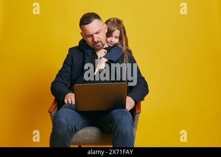 Thoughtful father using laptop and his beloved, little daughter pointing to screen against sunny-yellow background. Stock Photo