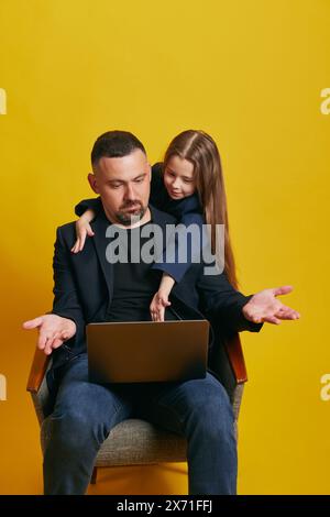 Little child, girl teaching his father how to use laptop pointing to screen of gadget against sunny-yellow background. Stock Photo
