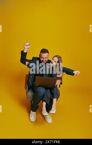 Business Partners. dad and his daughter collaborate over laptop, with joyful expression raising hands against sunny-yellow background. Stock Photo