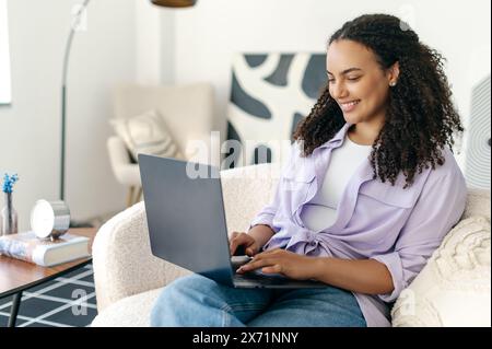 Gorgeous positive brazilian or hispanic young curly haired woman, freelancer or web designer, sitting on comfortable sofa, browsing internet via laptop computer. Working from home concept Stock Photo