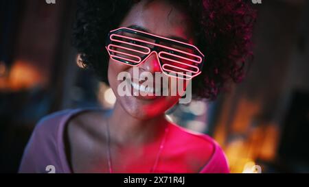 Close Up Portrait of Multiethnic Young Black Latin Female with Afro Hair in Futuristic Neon Glowing Glasses, Having a Fun at Home, Smiling and Posing for Camera in Loft Apartment in the Evening. Stock Photo