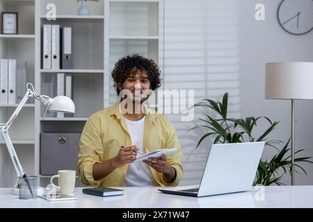 A cheerful man participates in an online course, taking notes while watching a webinar on a laptop in a modern home office. Stock Photo