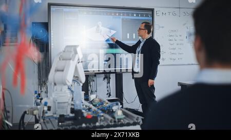 Male Professor Showing at the Big Screen During the Lesson. Diverse Group of Students Working Together on Modification of Functions of Technology Robot Arm for Engineering Project at University Stock Photo