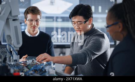 Male Student Holding a Tablet And Pointing at Robotic Hand While Working on Project with Asian Classmate and Black Teacher. Modern Technologies, Science and Developing at University Concept. Stock Photo