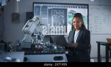 Portrait of Smiling Black Developer Using Laptop while Working with Robotic Arm in Modern University Classroom. Woman Looking at Camera. Success and Programming Concept. Medium Shot Stock Photo