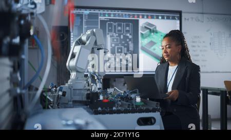 Black Female Developer Controls Robot Hand with Laptop, while Writing Code at Computer Science Research Laboratory. High-Tech Science and Programming at Modern Start Up Lab Concept. Stock Photo