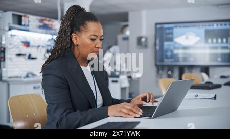 At the Factory: Black Automation Engineer Uses Laptop for Programming Robotic Arm while Sitting at Table and then Going to Robot. New Era in Automatic Manufacturing Industry Concept. Stock Photo