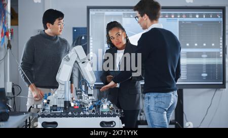 Black Teacher with Tablet and Two Diverse Students Analyse and Discuss Futuristic Robotic Arm During the Lesson. Lecturer Helping Engineering Majors with Project. Computer Science Concept. Stock Photo