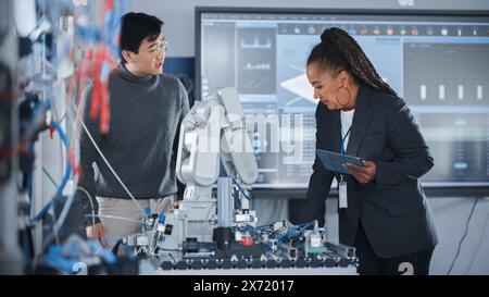 Teacher and Engineer Student Meeting in Robotic Research Laboratory: They Gathered Around Table, Talking, Using Tablet and Analysing Design of Industrial Robot Arm. Developing Concept. Medium Shot Stock Photo