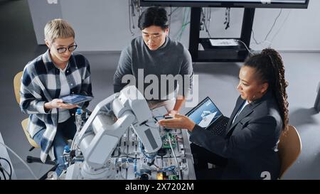 Black Teacher Explaining Information to Students Gathering Around Table with Robot Limb. People Holding Devices with Apps and Discussing It at University. Education Concept. High Angle View Stock Photo