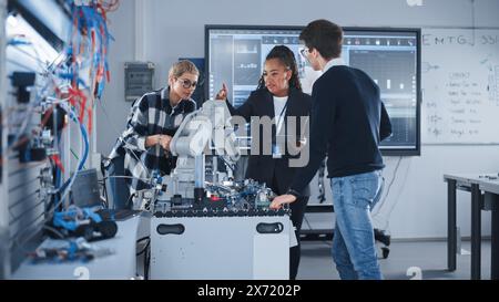 In Robotics Development Laboratory: Black Female Teacher and Two Students Work With Prototype of Robotic Hand. Young Student Telling Something with Smile and Brainstorming with Her Team. Stock Photo