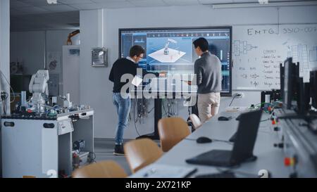 Two Diverse Male Students Standing Near Big Screen and Developing Program for Motion of Robotic Arm. Education and Innovative Equipment in Modern University of Technology Concept. Stock Photo