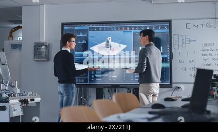 Diverse Team of Two Students Standing Near Big Screen with Tablet and Laptop While Discussing Programming and Technologies. Education and Studying in Modern University of Technology Concept. Stock Photo