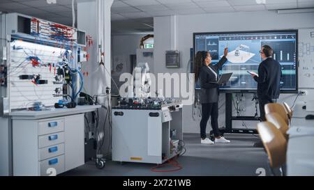 Female Black Developer Looking at Big Screen and Telling her Ideas to Male Colleague. Robotic Arm Moving in Background. Science and Innovative Technologies on Modern Factory Concept. Stock Photo