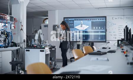 Black Female Automation Engineer Holding Laptop and Moving Bionic Arm With Written Code at Futuristic Factory. Modern Equipment and New Era in Computer Science and Robotics Industry Concept. Stock Photo