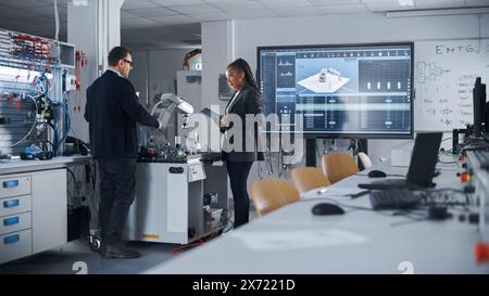 Diverse Colleagues Standing Near Table With Robot Arm And Discussing Complicated High-Tech Project While Using Digital Tablet. Bright Facility, Creative People Working in Start Up Concept. Stock Photo