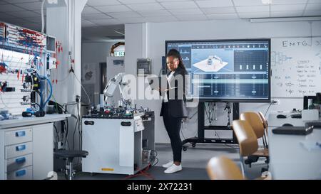 Black Female Automation Engineer Holding Laptop and Moving Bionic Arm With Written Code at Futuristic Factory. Modern Equipment and New Era in Computer Science and Robotics Industry Concept. Stock Photo