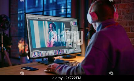 Young Creative Man in Headphones Using Computer in Stylish Loft Apartment in the Evening. Graphic Designer Working from Home, Editing Fashion Photo for Online Store. Urban City View from Big Window. Stock Photo