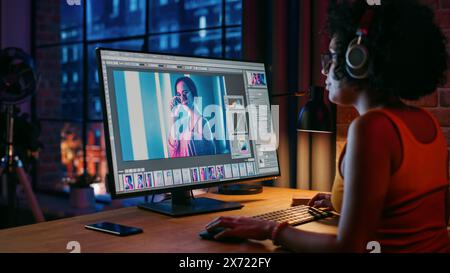 Young Creative Female in Headphones Using Computer in Stylish Loft Apartment in the Evening. Graphic Designer Working from Home, Editing Fashion Photo for Online Store. Urban View from Big Window. Stock Photo