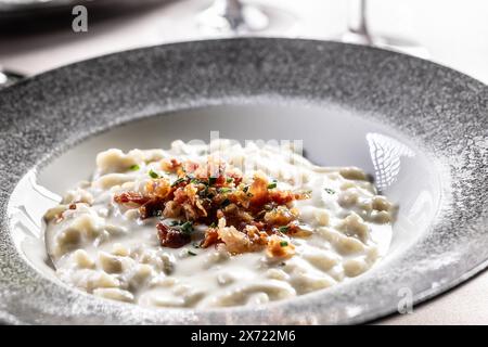 Halusky as traditional Slovak potato gnocchi with sheep cheese bryndza, fried bacon and chives served in a modern plate and on a festively laid restau Stock Photo