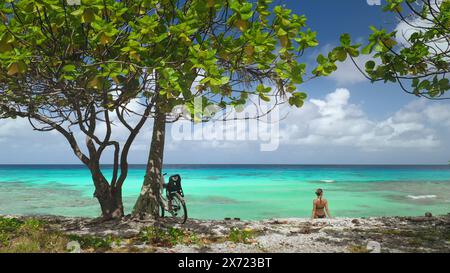 Tourist woman sitting on azure sea beach, framed by lush greenery and a tree with a bicycle leaning against, tranquility of a tropical shoreline on a clear day. Travel exotic background Stock Photo
