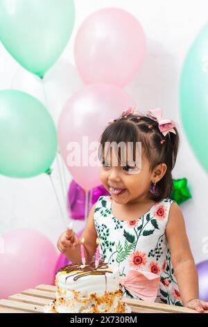 little latin brunette girl smearing her finger with cake to try it, surrounded by colorful balloons Stock Photo