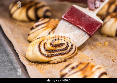 Person glazing raw cinnamon rolls with a pastry brush and preparing them for baking Stock Photo
