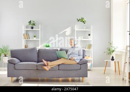 Senior woman using laptop and smiling while sitting on couch in at home in headphones. Stock Photo