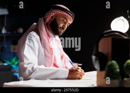 Side-view of a focused male entrepreneur using his laptop while seated at a desk. Arab guy focuses on his studies while using his laptop to explore the internet while surrounded by a notepad. Stock Photo