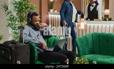 Tourist watches tv show in lounge area at luxuious hotel, waiting for check in process and killing time with movie on laptop. Cheerful person using computer in reception hall after travelling. Stock Photo