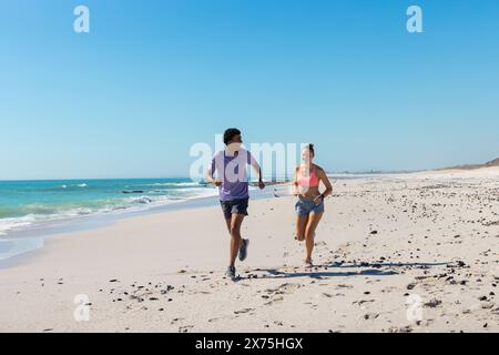 At beach, diverse couple wearing summer clothes, running on sunny beach Stock Photo