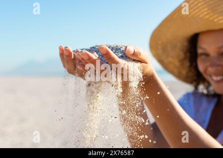 At beach, biracial young woman wearing hat playing with sand Stock Photo