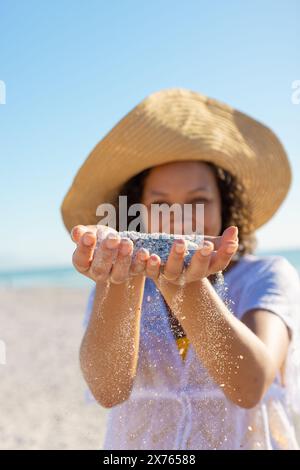 At beach, biracial young woman wearing sun hat holding sand Stock Photo