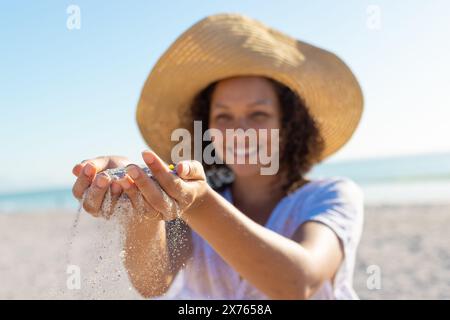 At beach, young biracial woman playing with sand, wearing hat Stock Photo