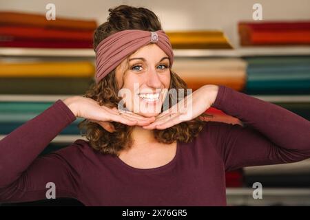 A playful fashion designer showcases her cheerful personality, framing her face with hands while wearing a trendy headband in her vibrant textile stud Stock Photo