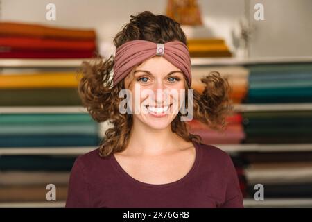A playful fashion designer with curly hair and sparkling eyes showcases a mauve headband, standing in front of colorful fabric rolls in her workshop Stock Photo
