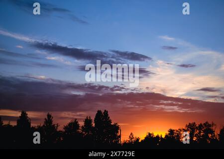 The morning glow illuminates the clouds on a beautiful colorful sky above the silhouettes of trees and a pillar at dawn Stock Photo