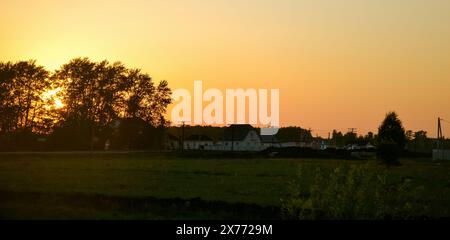 The setting sun behind the silhouettes of trees and the yellow evening sky over the houses in the village Stock Photo
