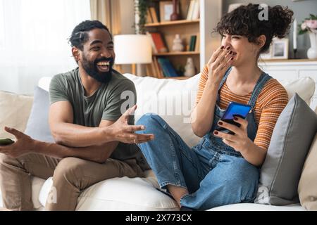 Young happy couple, simple living. African American man and caucasian woman, hipsters and millennial enjoying and laughing at home, sitting on sofa in Stock Photo