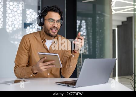 Man wearing headphones attending an online webinar while taking notes. He is sitting at a modern office desk with a laptop and notebook. Stock Photo